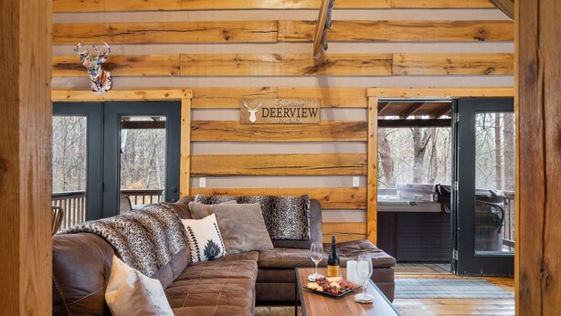 Interno di una baita in legno con un divano in pelle marrone, un tavolino da caffè con vino e snack, e un cartello appeso al muro con la scritta "Deerview".
