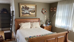 A bedroom with a wooden bed, a nightstand, a dresser, a painting on the wall, and a window with curtains. - The Coffey House (Choteau)