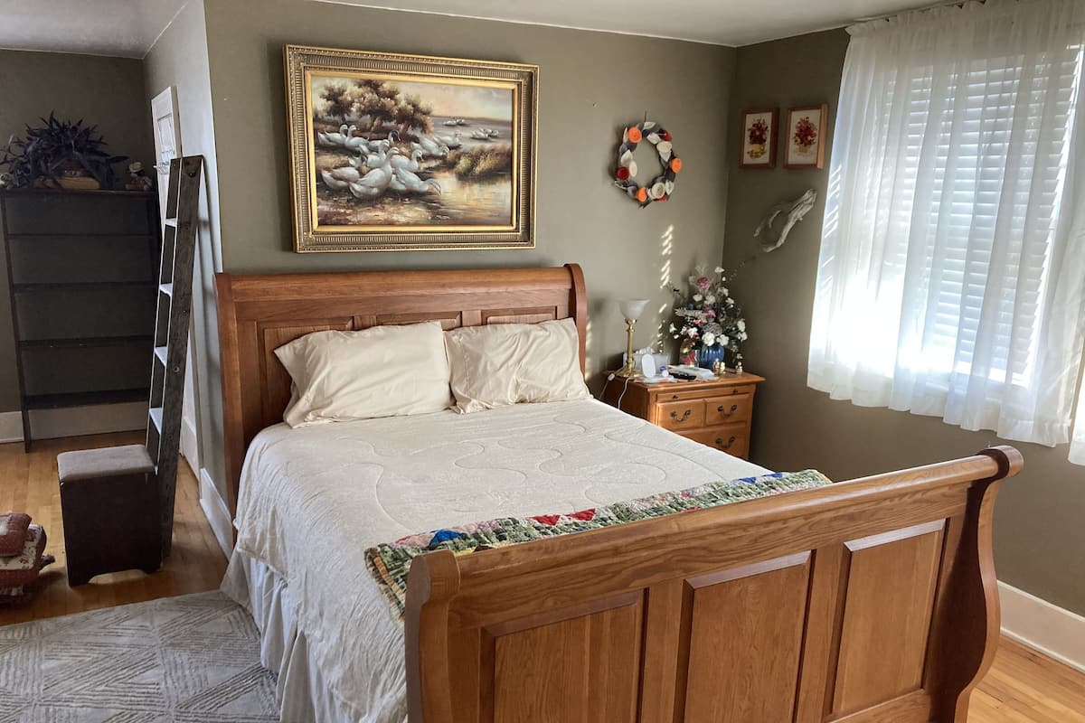 A bedroom with a wooden bed, a nightstand, a dresser, a painting on the wall, and a window with curtains.