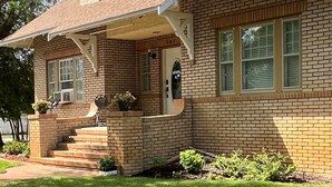 A two-story brick house with a front porch, a small garden, and a clear sky. - The Coffey House (Choteau)