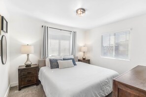 Ein Schlafzimmer mit einem großen Bett, zwei Nachttischen mit Lampen, einem Holznachtkästchen und zwei Fenstern mit Jalousien.