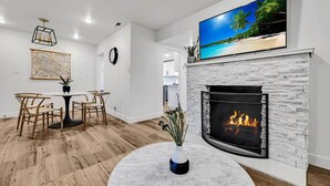 Ein modernes Wohnzimmer mit Kamin, einem Essbereich mit Holzsesseln und einem großen Flachbildfernseher, der über dem Kamin angebracht ist und eine Strandansicht zeigt.