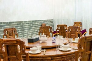 Una mesa de madera redonda con sillas, un calentador de agua, tazas y platillos, todo sobre un fondo de pared de ladrillo y cortinas blancas.