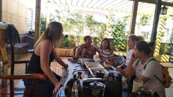A group of people around a table with various items, including a kettle and containers, in an outdoor setting with a wooden structure overhead.