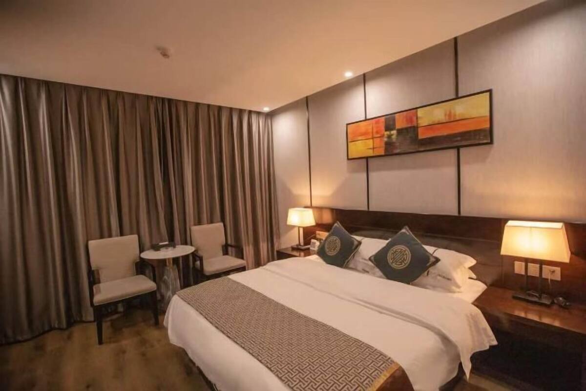 A hotel room with a large bed, two bedside tables with lamps, a small round table with a chair, and a large window with curtains.