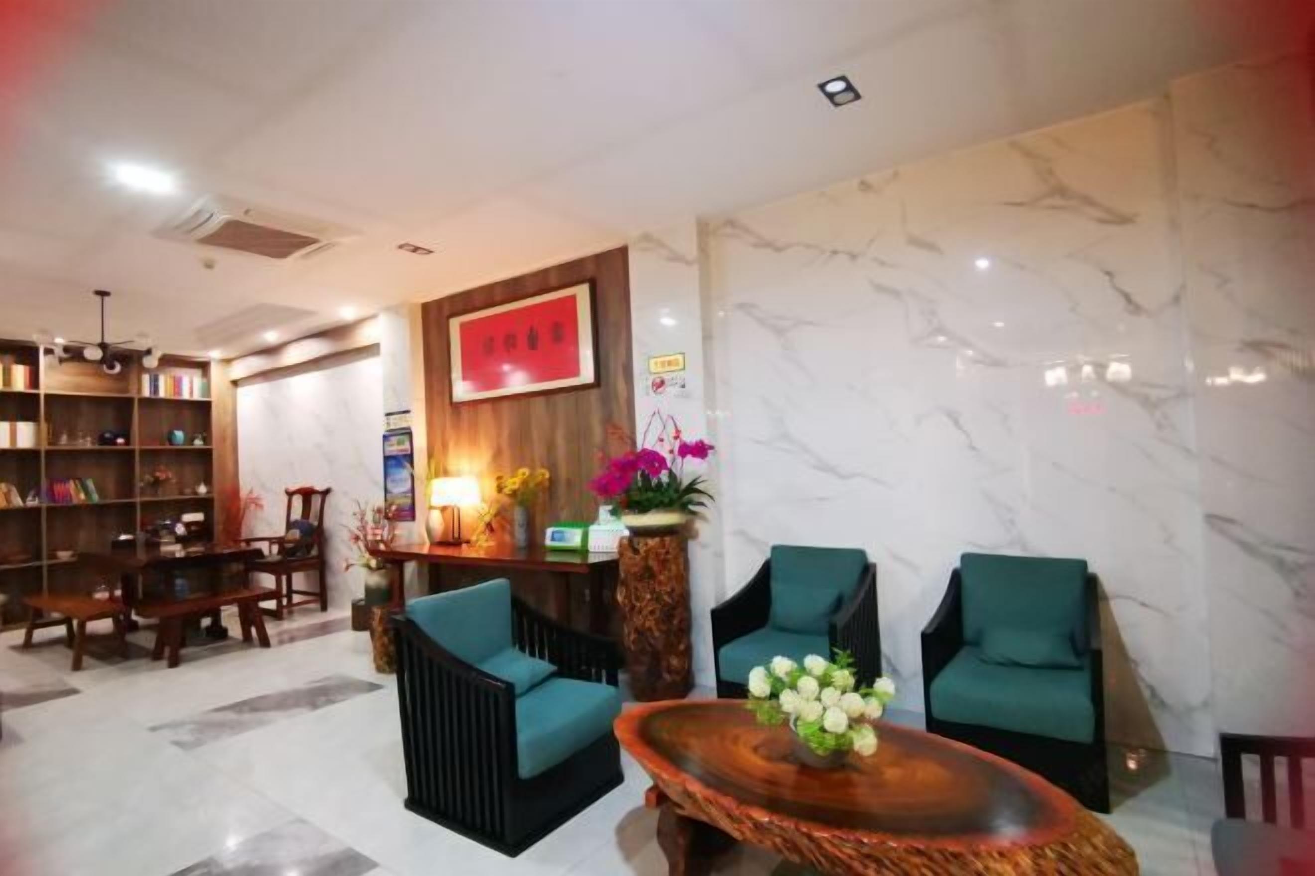 Um lobby moderno com uma mesa de recepção de madeira, duas poltronas verdes, uma mesa de centro redonda de madeira e uma estante repleta de livros.