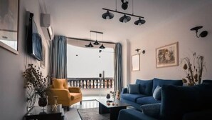 A modern living room with a blue sofa, a yellow armchair, and a coffee table. There is a large window with curtains, a flat-screen TV mounted on the wall, and a ceiling light fixture. - Pyramids Bay Apartment the best place to view the Giza Pyramids (Giza Governorate)