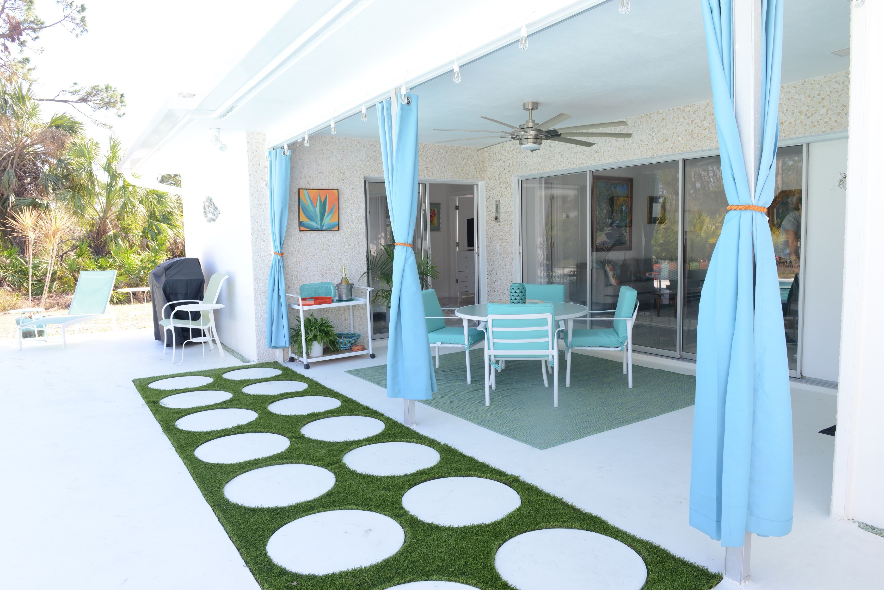 Een terras buiten met een witte tafel en stoelen, een blauwe parasol en een gebied met kunstgras in het groen met witte cirkels.