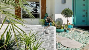 Une maison à l’extérieur blanc, avec une porte turquoise et un petit jardin agrémenté de diverses plantes en pot et d’un chemin de pierres.