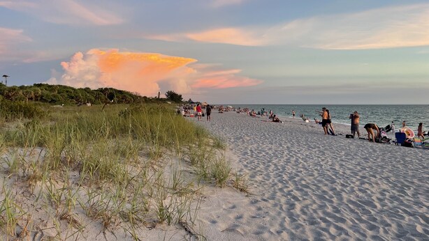 Une plage où des gens se détendent et prennent des bains de soleil, des dunes herbeuses et un ciel nuageux.