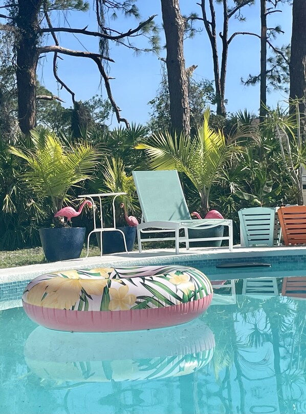 Un área de piscina con sillones reclinables, una estatua de flamenco y plantas tropicales.