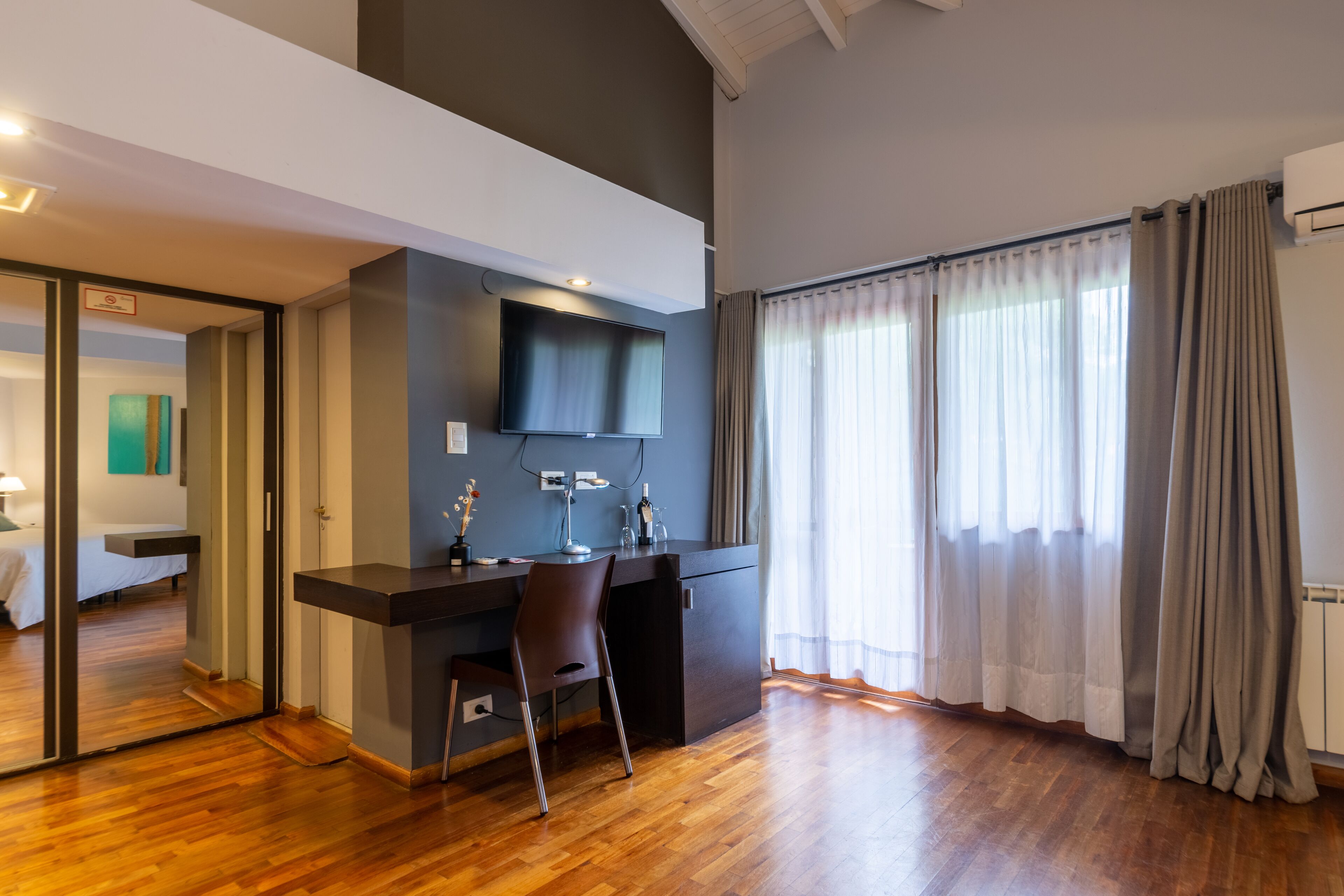 Une chambre moderne avec un plancher en bois, un bureau avec une chaise, un téléviseur fixé au mur et un lit à l’arrière-plan.
