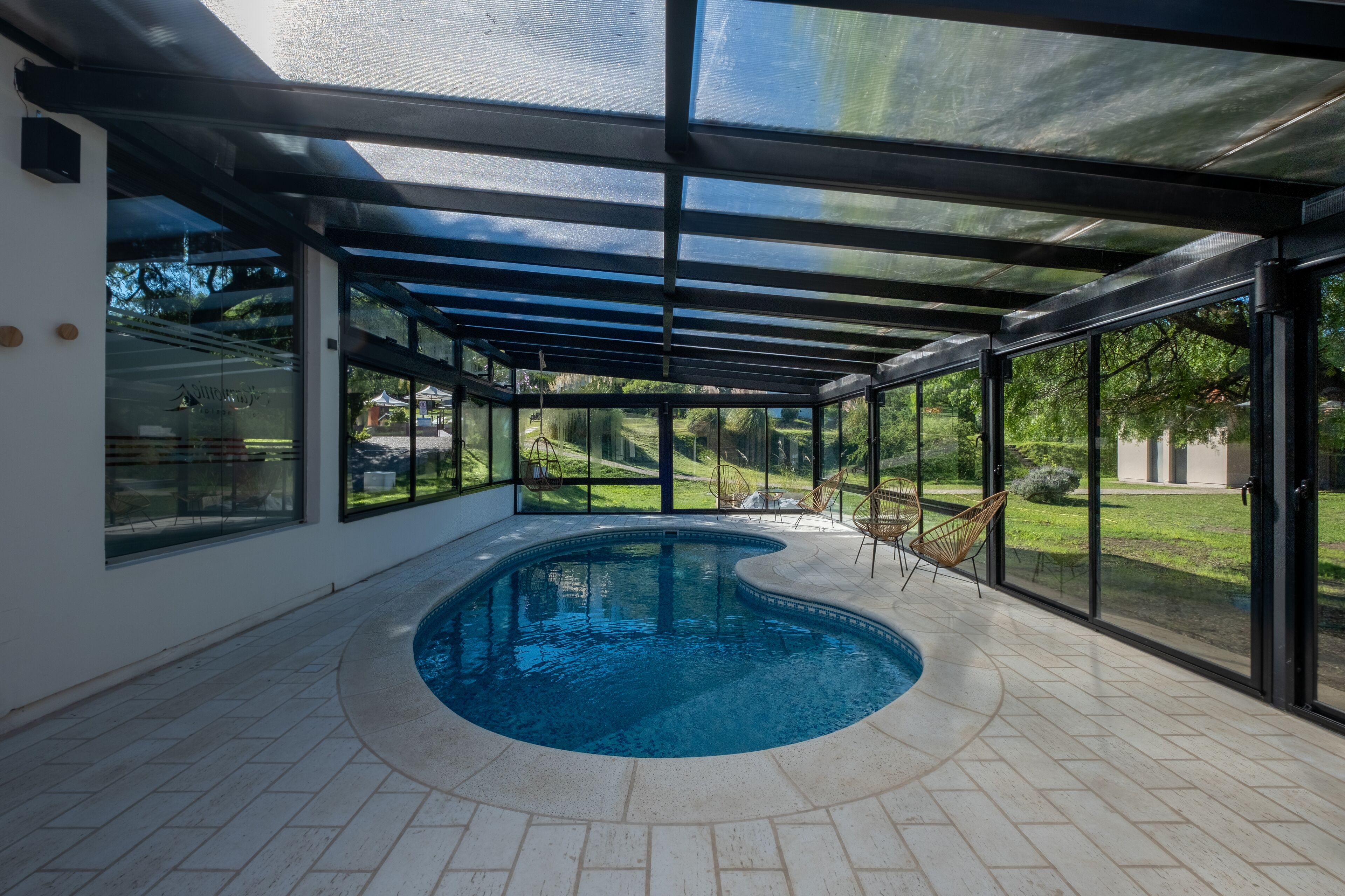 An indoor swimming pool with a glass enclosure, surrounded by large windows offering views of a green landscape.