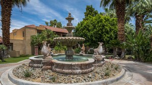 Una fuente de piedra circular con caños de agua, rodeada por un jardín ajardinado con palmeras y diversas plantas.