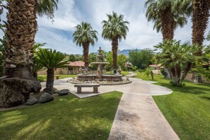 A landscaped garden with a central fountain, palm trees, and a paved walkway.