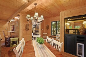 A wooden cabin interior with a dining table, chairs, a chandelier, a kitchen area, and a large window. - Pet Friendly, Fenced Yard, 3 Bedroom Cabin, Hot Tub, 3 Mi. to Beech Ski Resort!  (Beech Mountain)