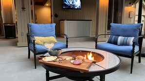 En mysig utomhusplats med två blå stolar, ett runt bord med en eldstad och en tv som är monterad på väggen.