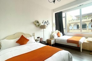 A hotel room with two beds, a window with a view of a building, a lamp, and a wall with photos.
