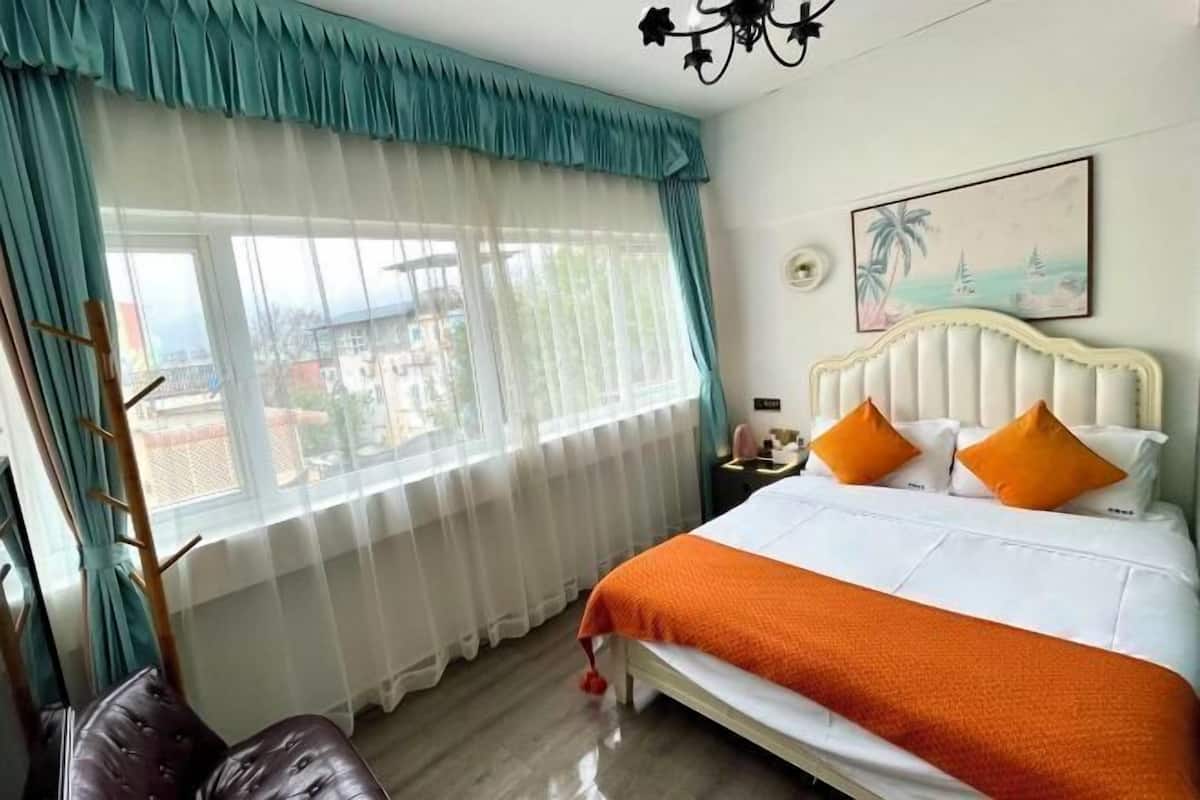 Um quarto com cama, janela com cortinas, um quadro na parede e mesa de cabeceira com abajur.