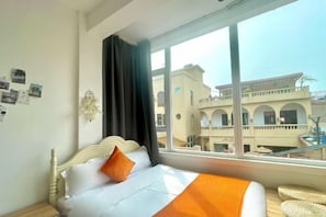 A hotel room with a bed, a window with a view of buildings, and a wall with pictures.