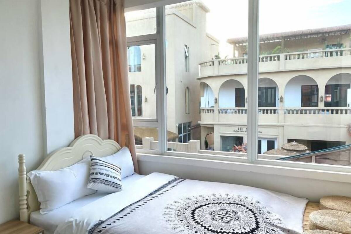 Uma cama com cabeceira branca, manta com padrão e vista para um edifício com varandas através de uma grande janela.