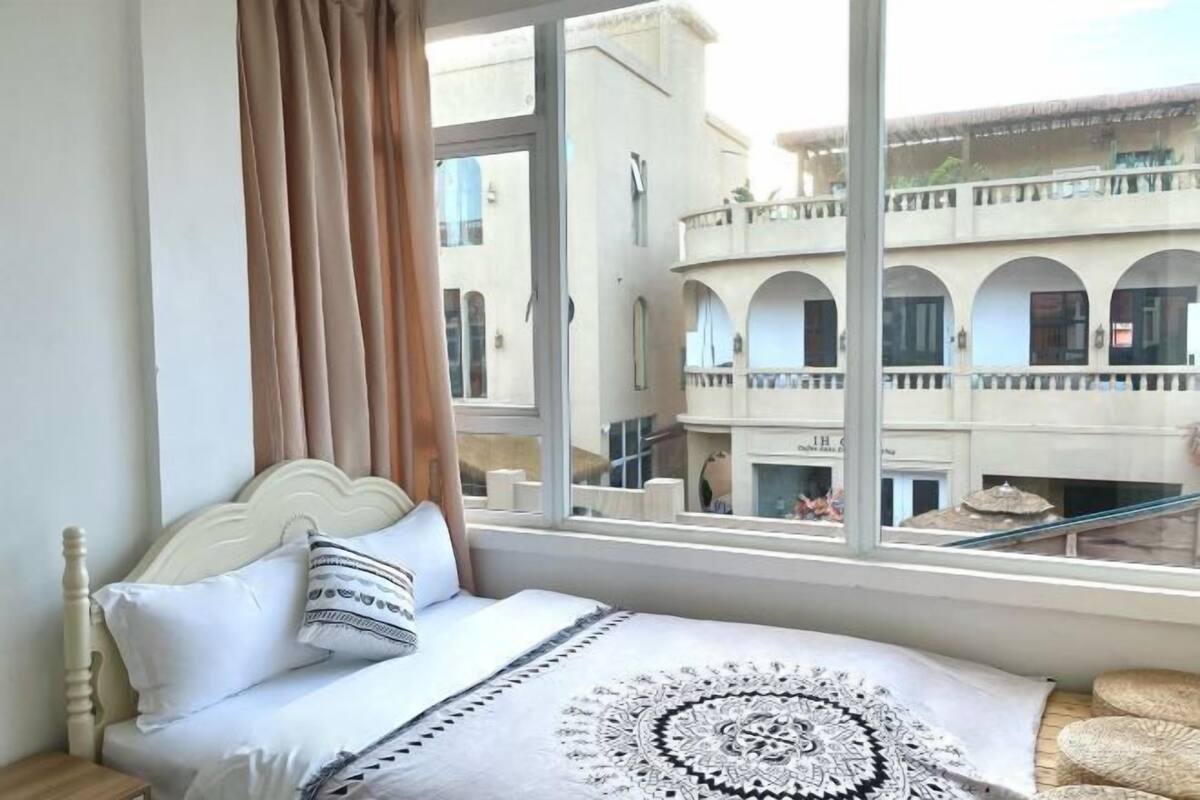 Quarto com cama, cortinas, janela e vista para um edifício com varandas.