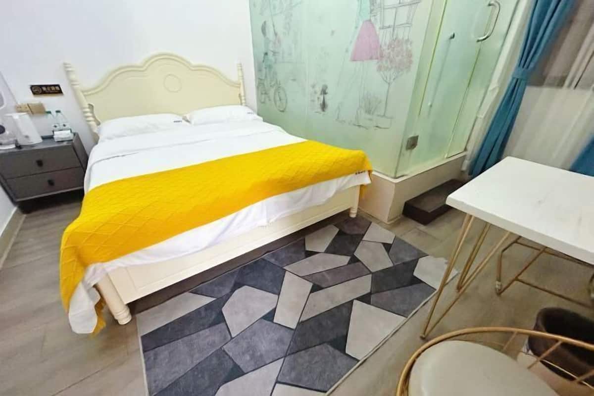Cama com cobertor amarelo, cabeceira branca e tapete estampado. Há uma pequena mesa e uma cadeira à direita.