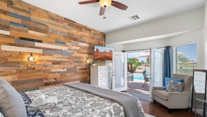 A bedroom with a wooden wall, a bed with patterned bedding, a ceiling fan, and a view of an outdoor pool area. - Chandler Oasis Pool, Golf Views & Fire Pit (Chandler)