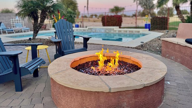 A fire pit with flames, surrounded by outdoor seating including chairs and a table, with a pool and palm trees in the background. - Chandler Oasis Pool, Golf Views & Fire Pit (Chandler)
