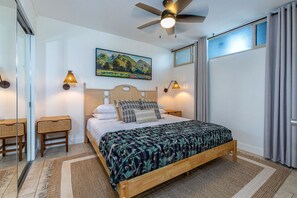Un dormitorio con cama, dos mesitas de noche, ventilador de techo, ventana con cortinas y un cuadro en la pared.