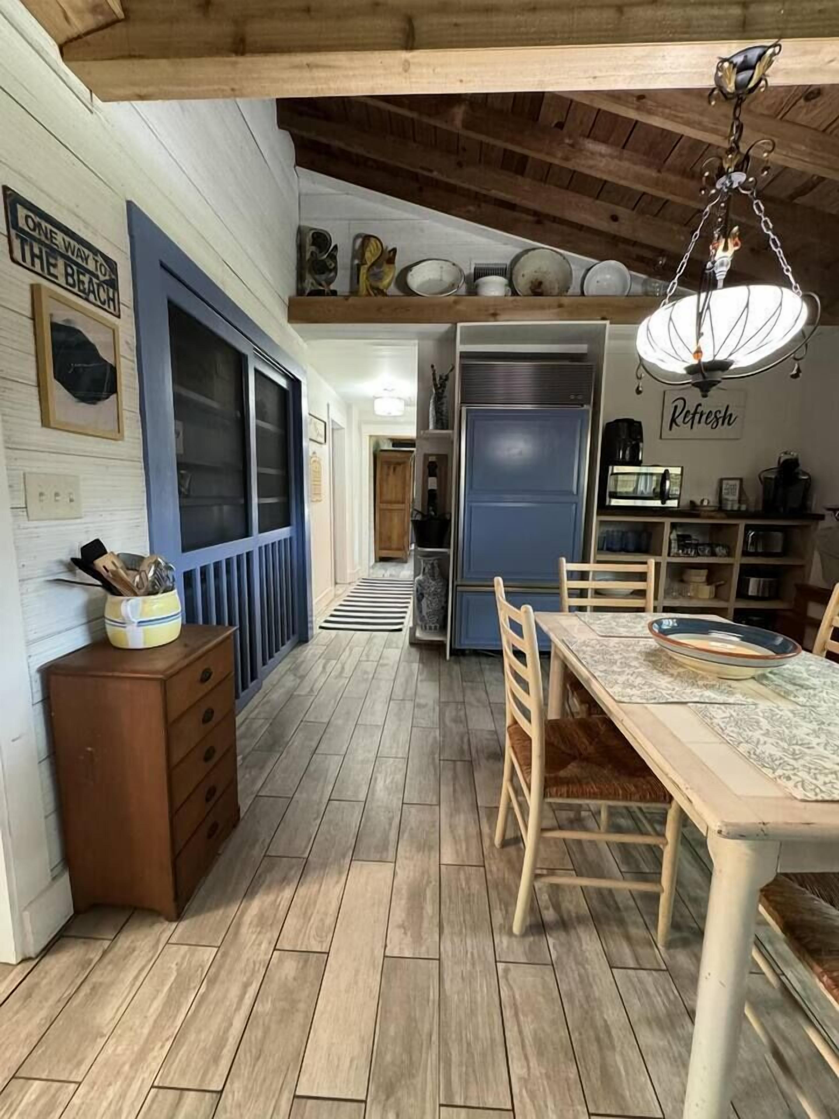 A kitchen with a wooden floor, a dining table with chairs, a refrigerator, and a hanging light fixture.
