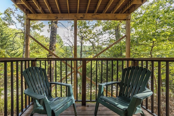 A wooden deck with two green Adirondack chairs, surrounded by trees.