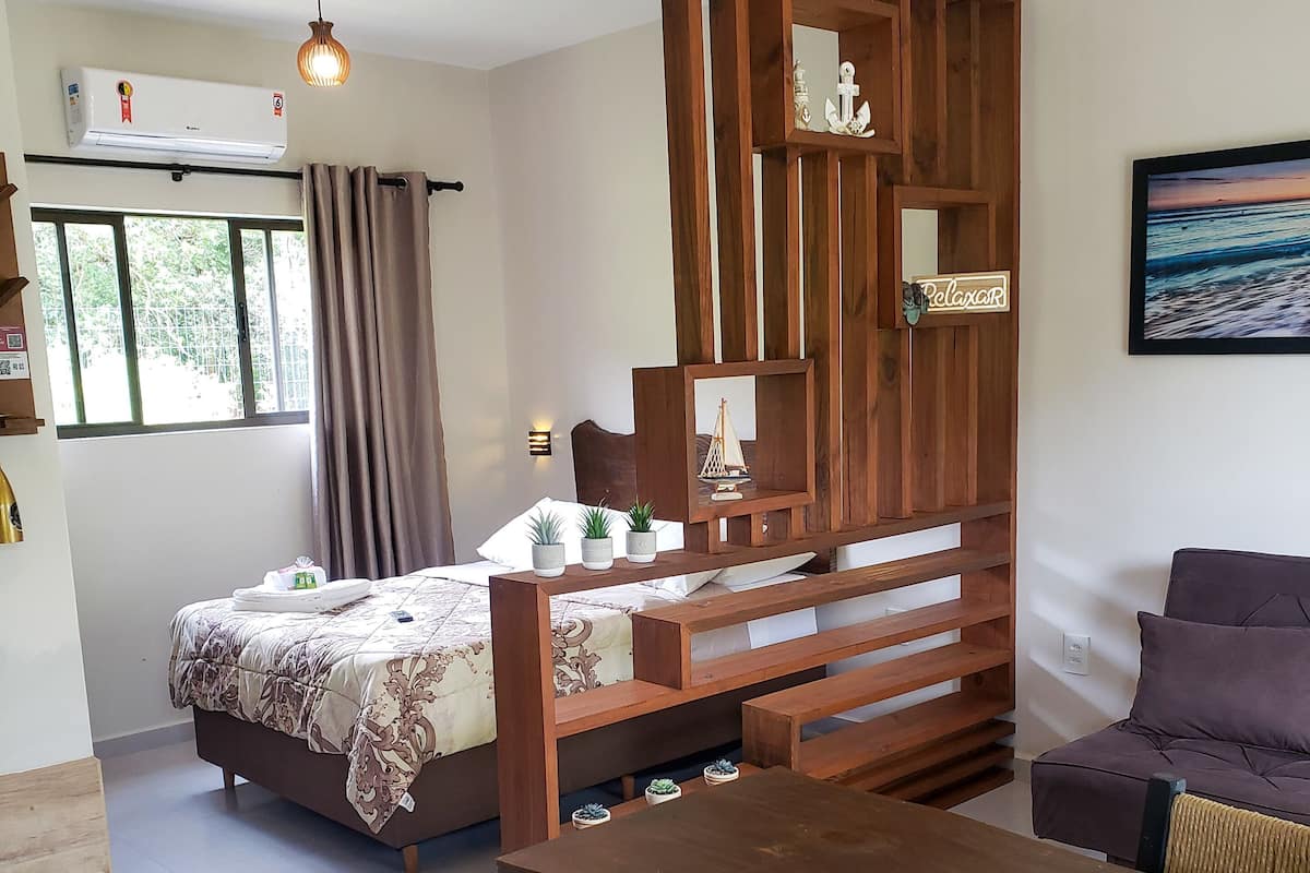 A bedroom with a bed, bedside tables, a wooden partition, a dining table with chairs, and a view of the outdoors.