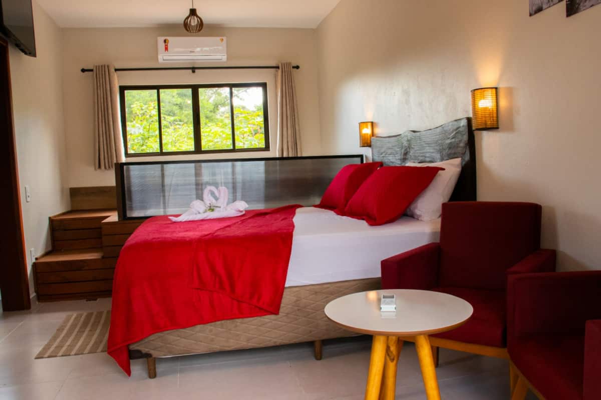 A hotel room with a bed, a small table, and a chair. There is a window with curtains, a staircase, and a wall-mounted air conditioner.