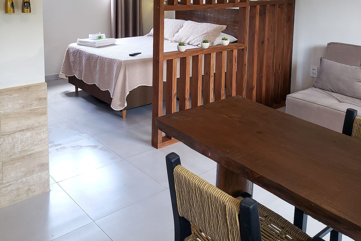 A room with a bed, a sofa, a dining table, a wooden partition, and a view of the outdoors.