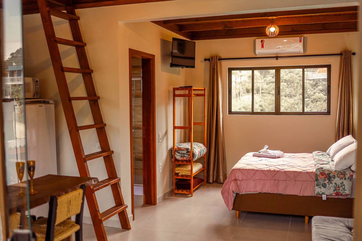 A bedroom with a bed, a wooden ladder, a TV, and a window with curtains.