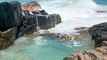 A rocky coastal area with clear turquoise water, waves crashing against the rocks, and a distant view of a hilly landscape with buildings.