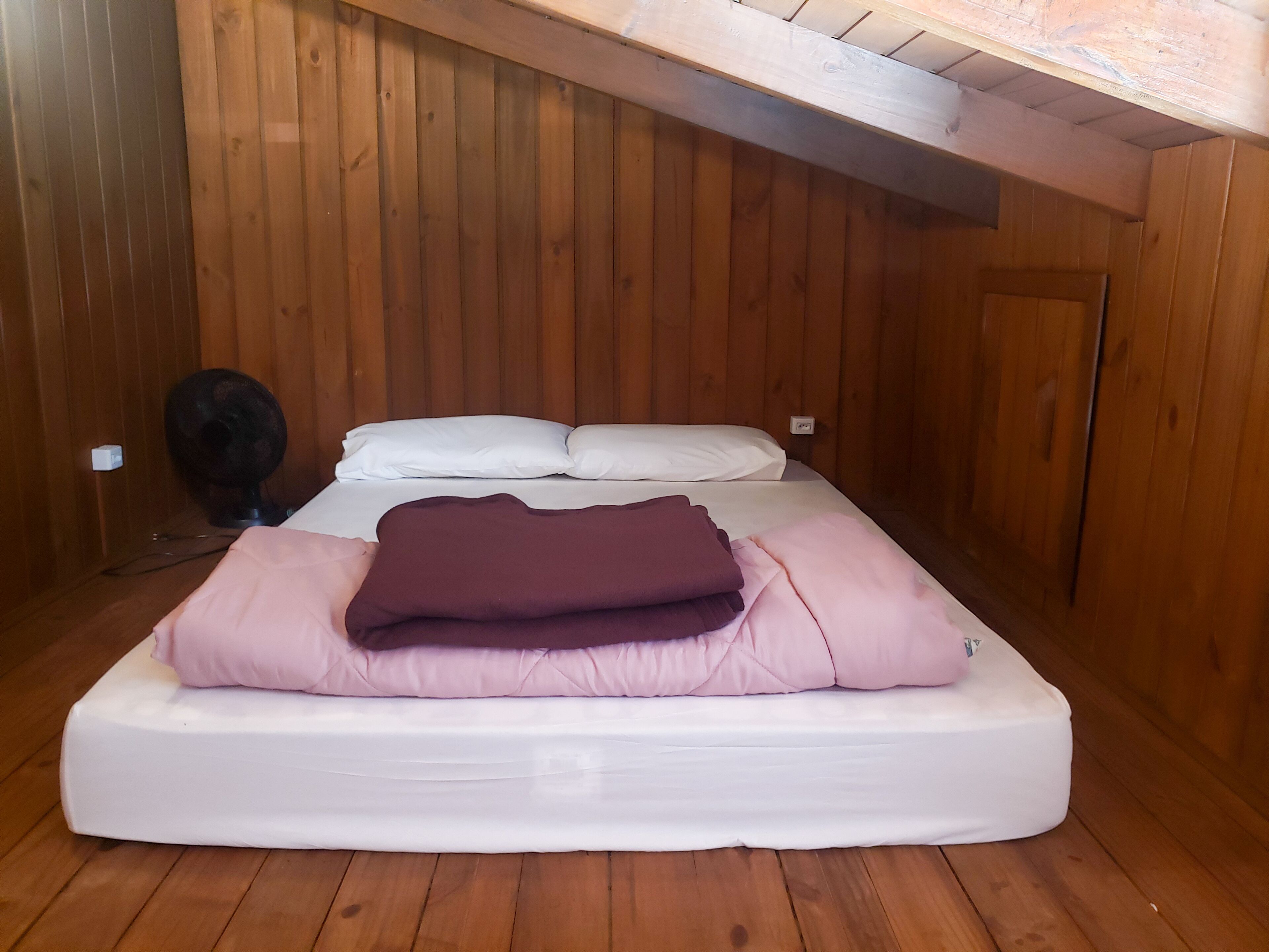 Una cama individual con ropa de cama blanca y rosa en una habitación con paredes de madera y techo inclinado.