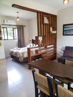 A room with a bed, a wooden bench, a dining table with chairs, and a wall-mounted shelf with decorative items. - Pousada Rosa Maria (Imbituba)