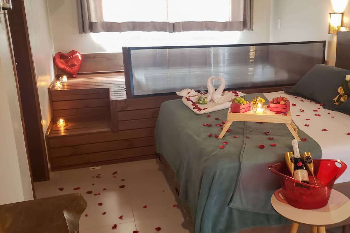 A hotel room with a bed, a bedside table with a candle, a heart-shaped balloon, a bouquet of flowers, a bottle of champagne, and rose petals on the floor.
