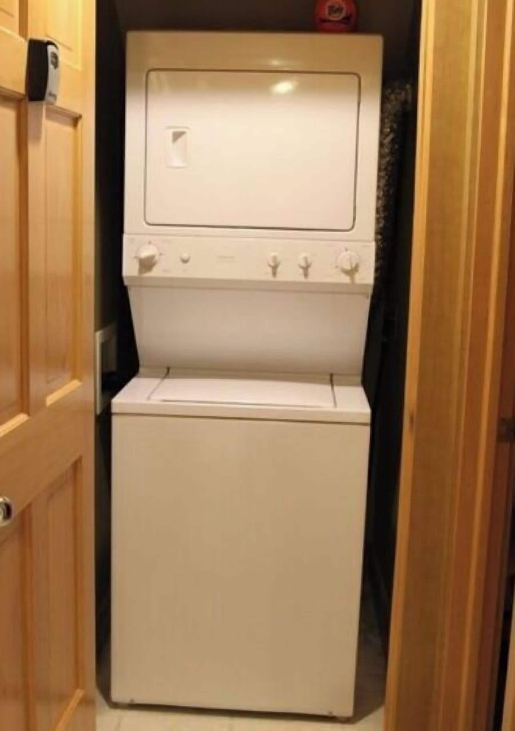 Lavadora blanca y compacta en un lavadero estrecho con puertas de madera y un ventanal pequeño.