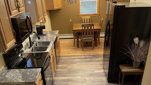 A kitchen with wooden cabinets, a granite countertop, a dining area with a wooden table and chairs, and a black refrigerator. - Rush Hollow Hideaway (Lake George)