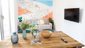 Una habitación con una mesa de madera, un florero azul, dos botellas de agua, un cuenco de madera, un control remoto y una pared con una ilustración de una cámara.