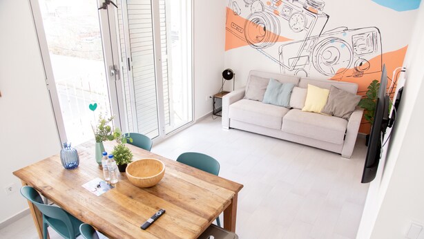 Un comedor con una mesa de madera, sillas color turquesa y un tazón. Hay un sofá con cojines y un mural en la pared.