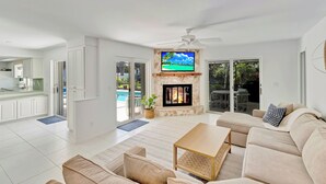 Un salón amplio con chimenea, un televisor montado encima y un ventanal grande con vistas a una piscina.