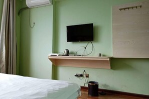 A room with a bed, a mounted television, a small shelf with a kettle and cups, and a wall-mounted air conditioner.