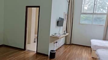 A hotel room with a wooden floor, a bed, a desk with a computer, a window, and a door.
