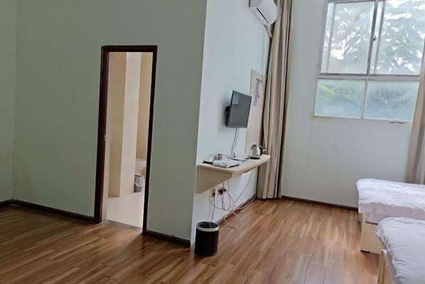 A hotel room with a wooden floor, a bed, a desk with a computer, a window, and a door.