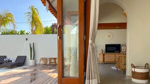 Um deck de madeira com cadeiras de descanso e um vaso de planta, que dá acesso a uma sala de estar com televisão e parede de tijolos.