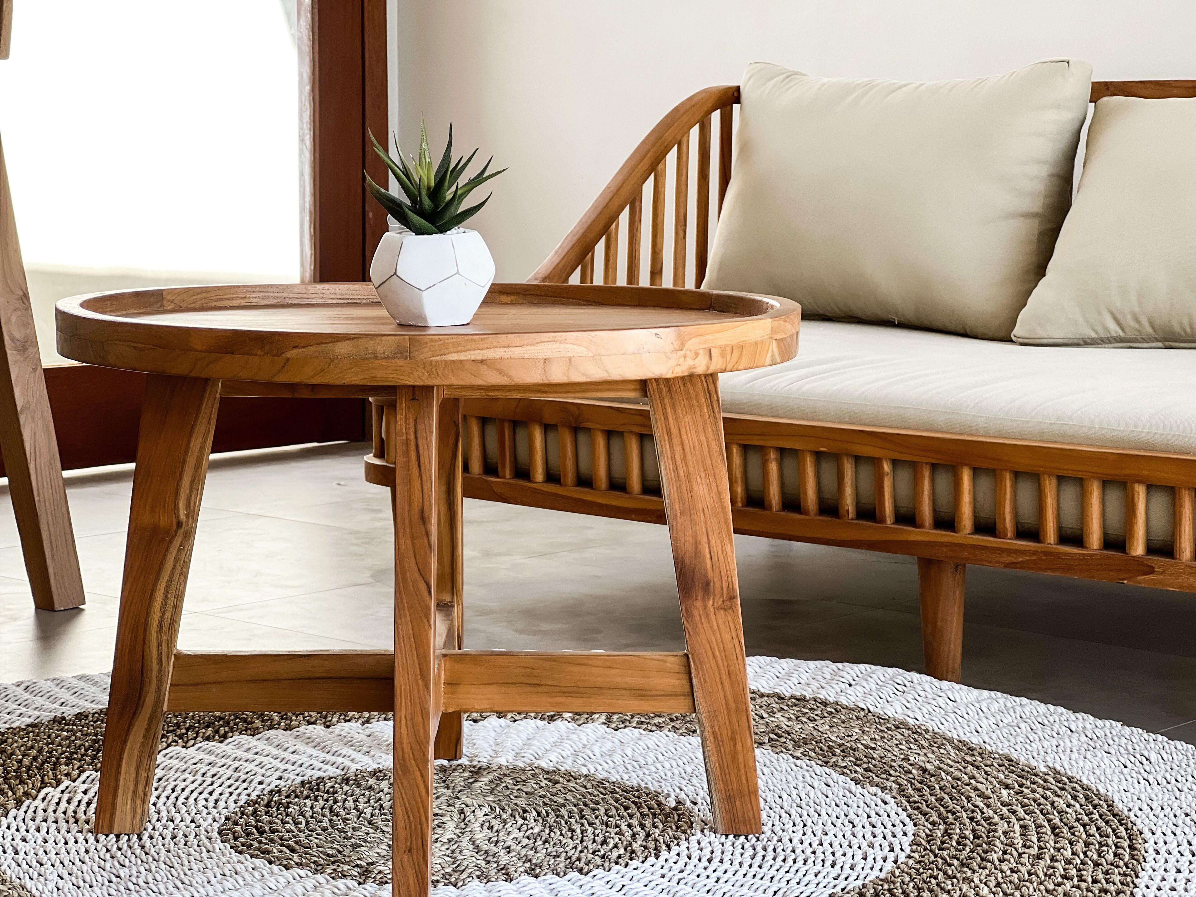 Une table ronde en bois, agrémentée d’une plante en pot, d’un tapis tissé et d’un banc en bois avec des coussins.